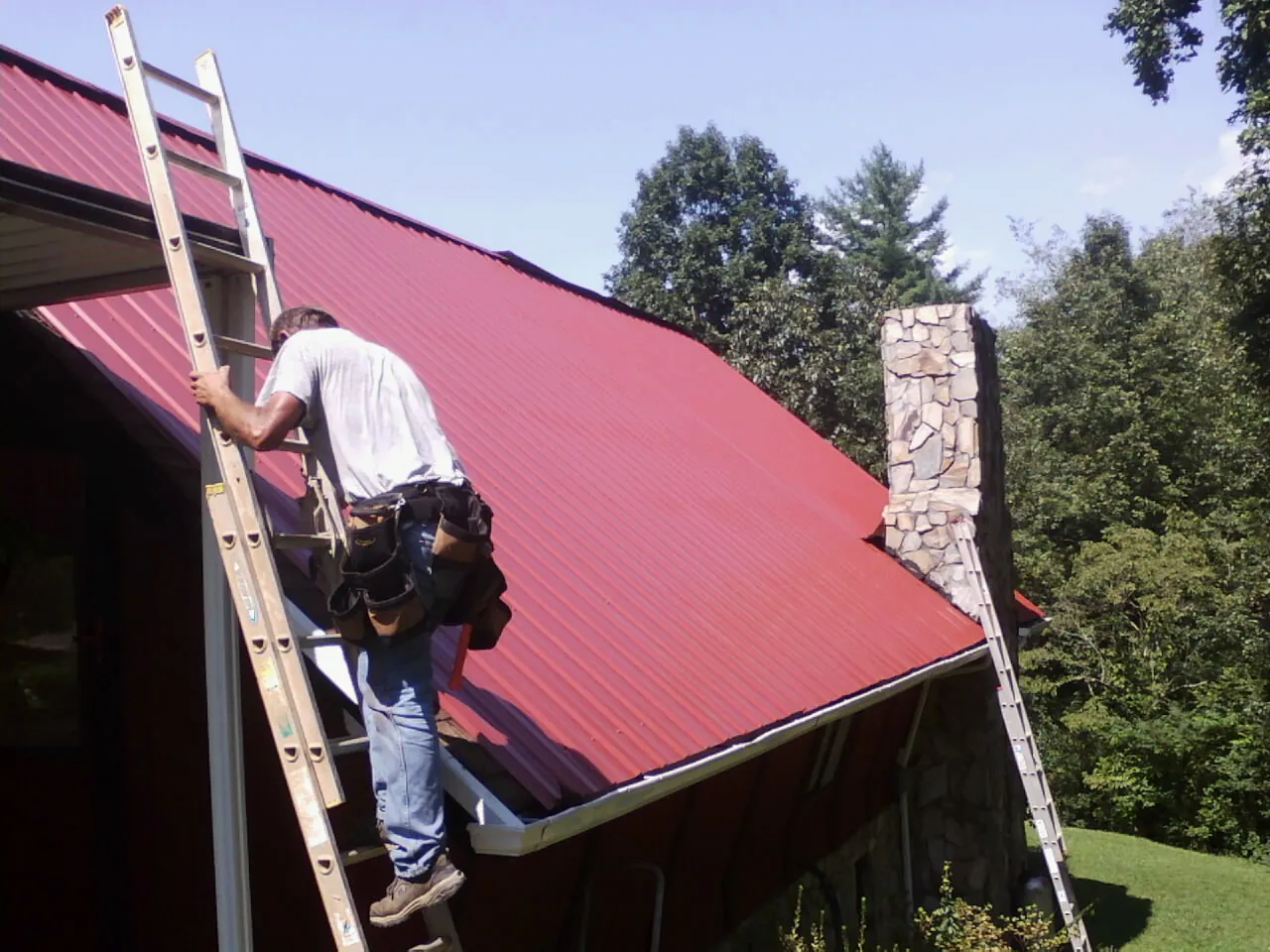 Roofer on a ladder beside a red metal roof and stone chimney — Soffit & Fascia Repair by Roofing Brothers in Holyoke